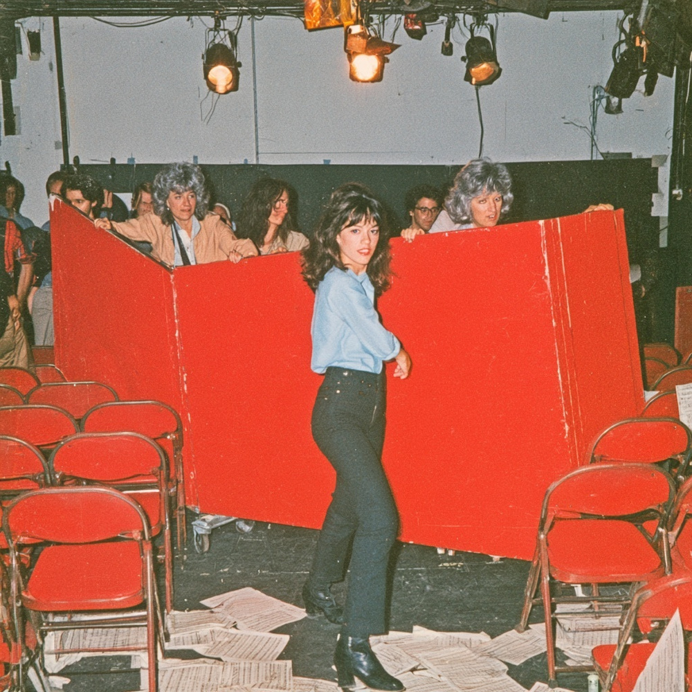 Woman in a blue shirt standing between red folding barricades in a small theater, with a gray-haired company and rows of red chairs.