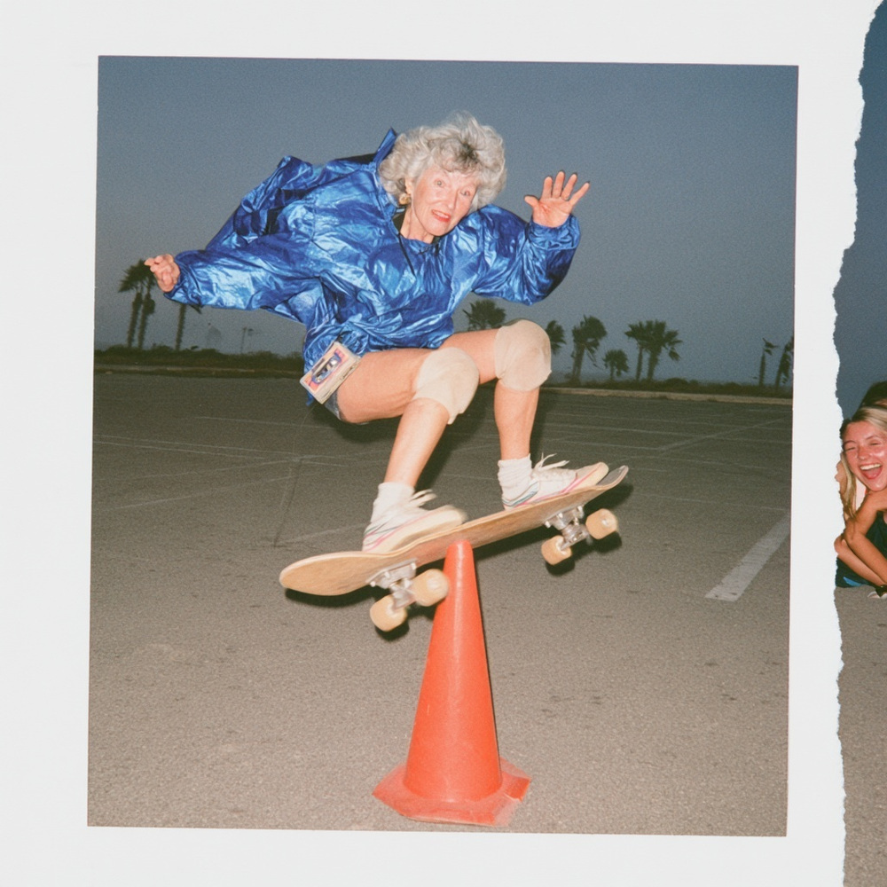 Silver-haired woman in a blue jacket airborne on a skateboard over an orange cone, with a laughing friend at the torn frame edge.