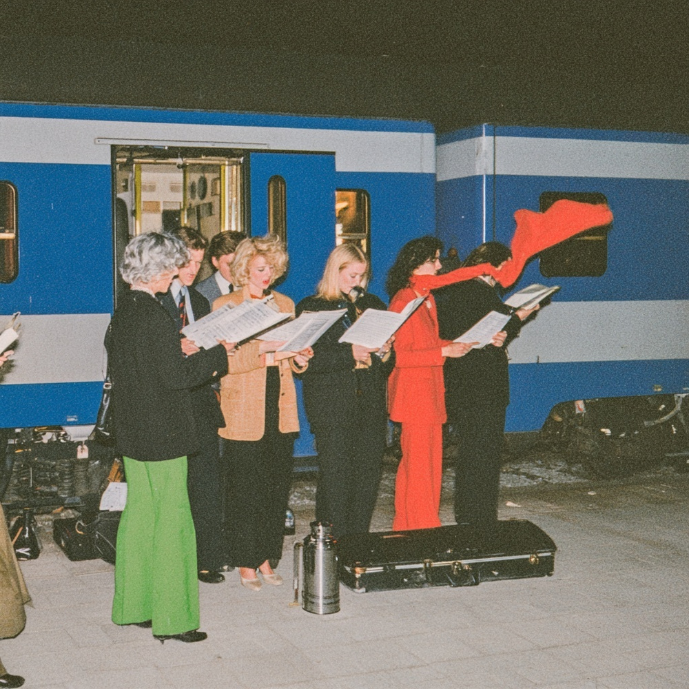 Mixed-age group singing from sheet music beside a blue train, red scarf flying above an instrument case and thermos.